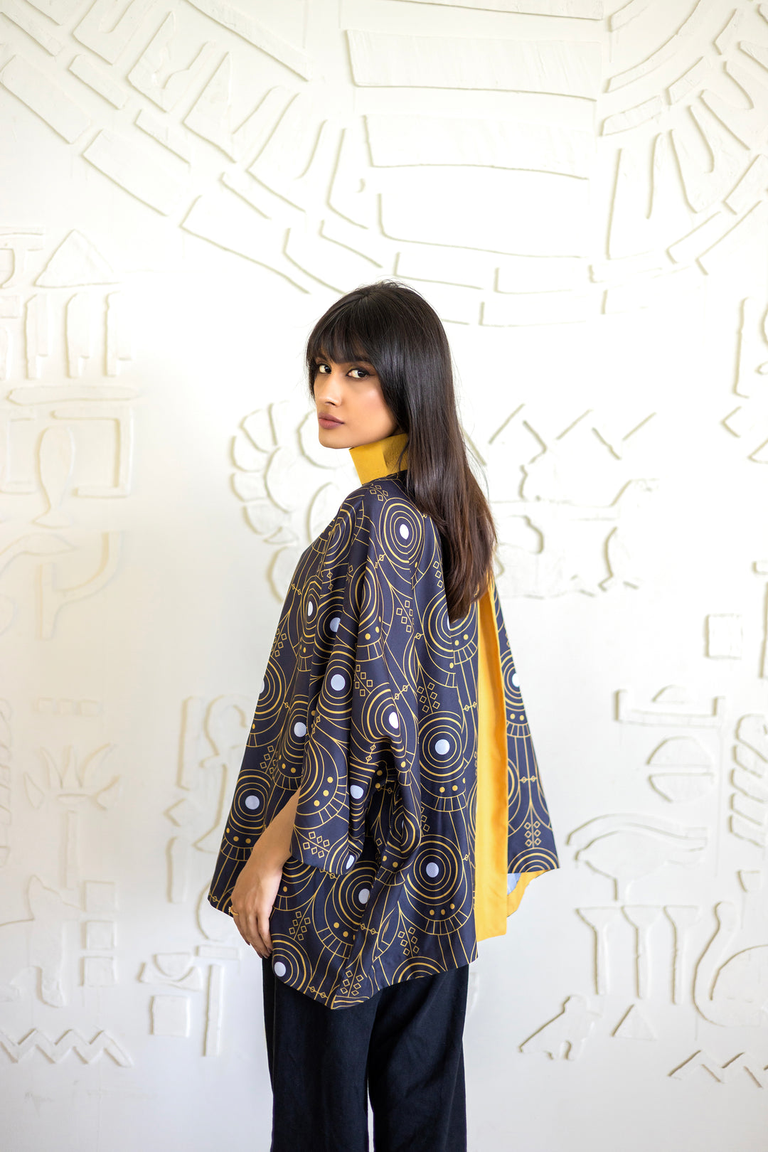 Woman wearing a patterned jacket over a yellow top against a white textured wall.