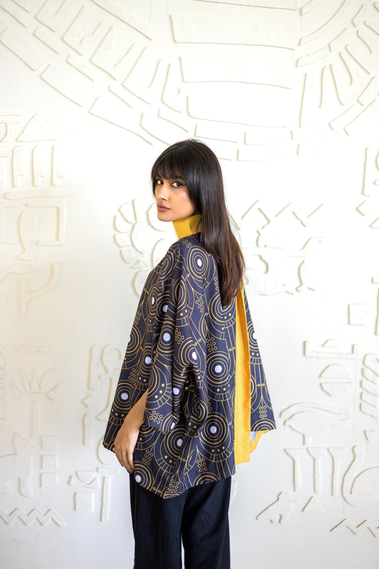 Woman wearing a patterned jacket over a yellow top against a white textured wall.