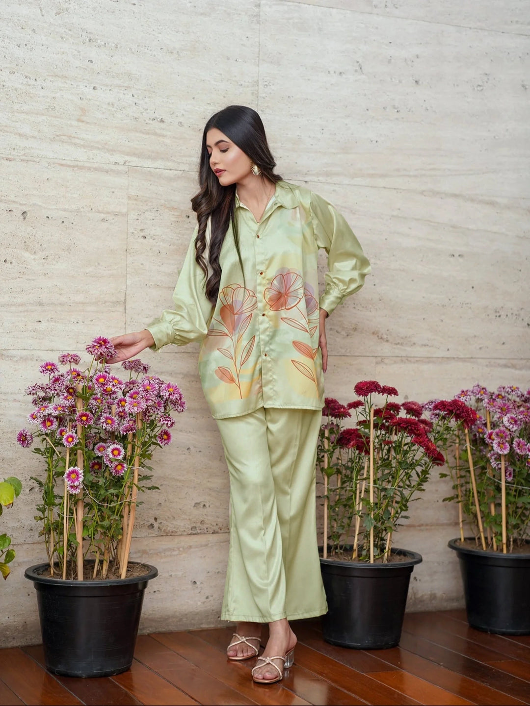 Woman in a light green floral outfit standing next to potted plants against a wooden wall.