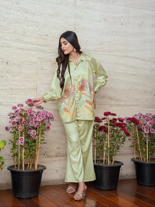 Woman in a light green floral outfit standing next to potted plants against a wooden wall.