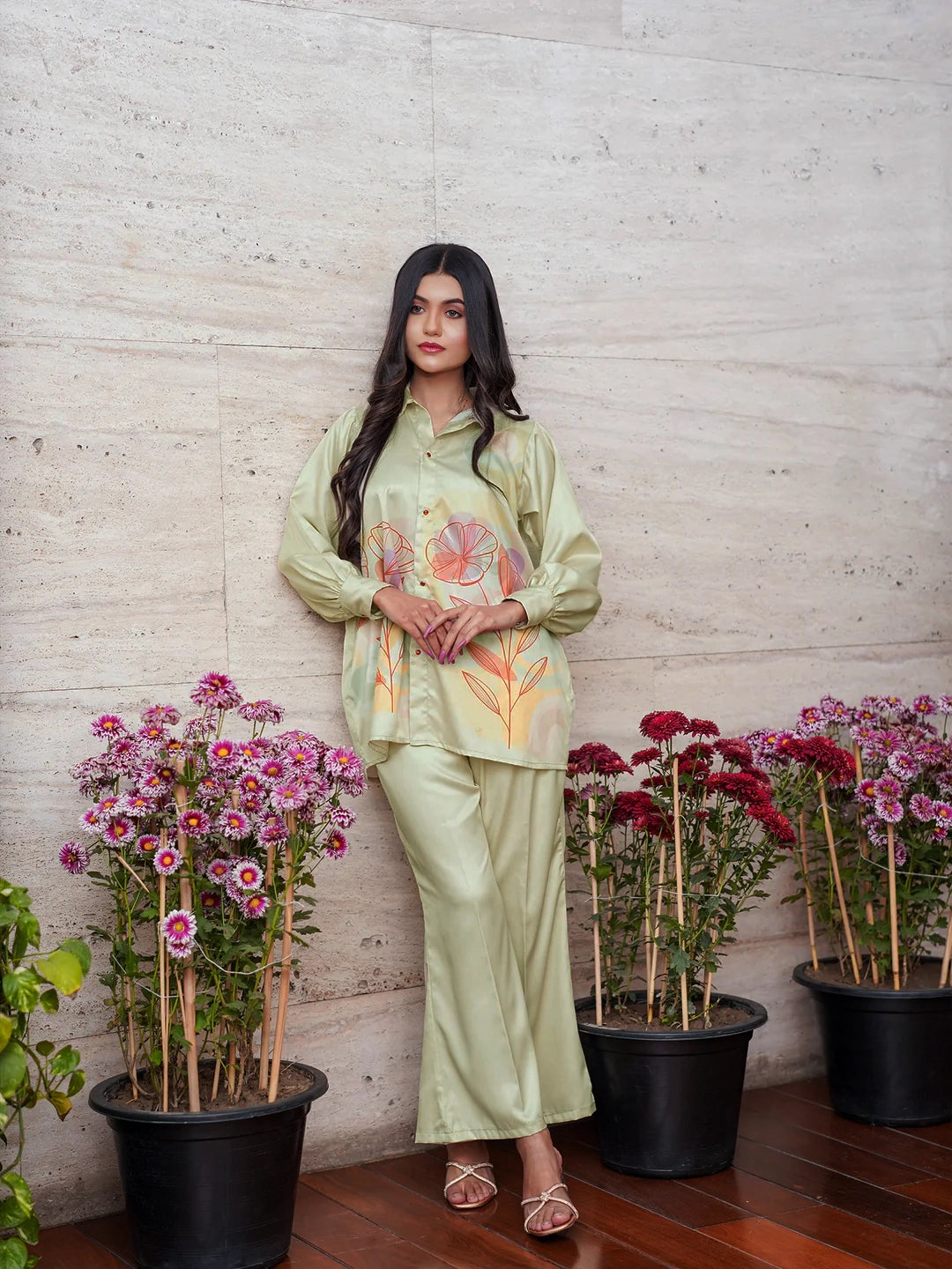Woman in a light green traditional outfit standing among potted plants against a textured wall.