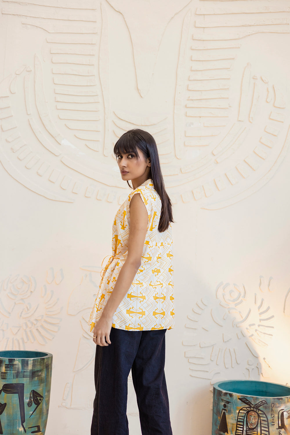 Woman wearing a sleeveless patterned top and dark pants against an abstract wall.