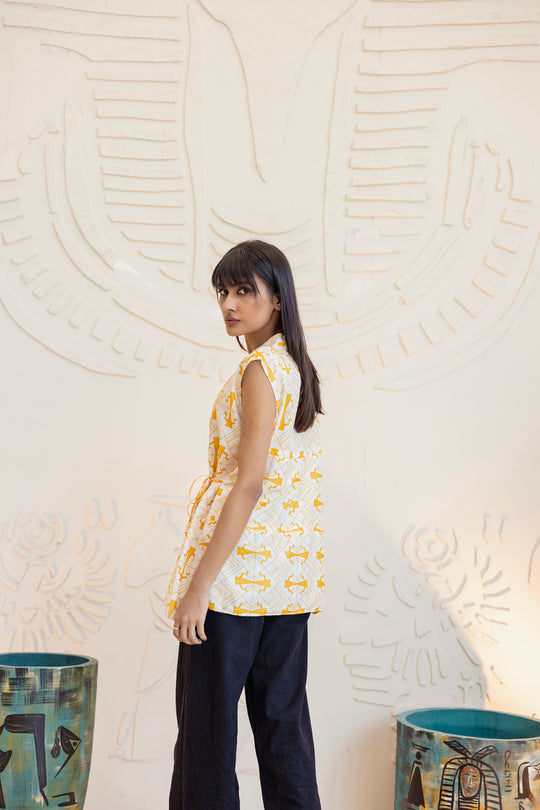 Woman wearing a sleeveless patterned top and dark pants against an abstract wall.