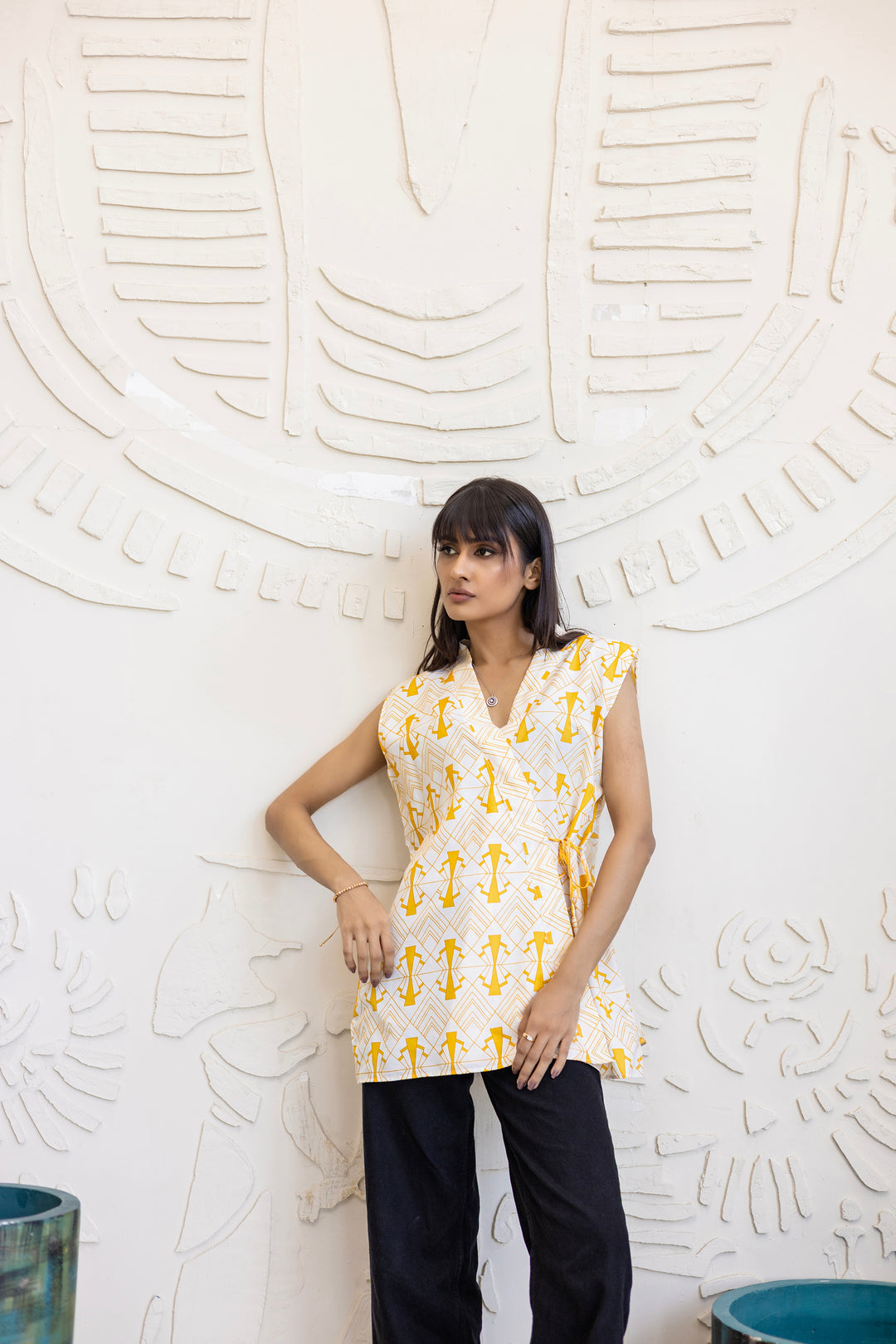Woman wearing a yellow and white patterned sleeveless top against a textured white wall.