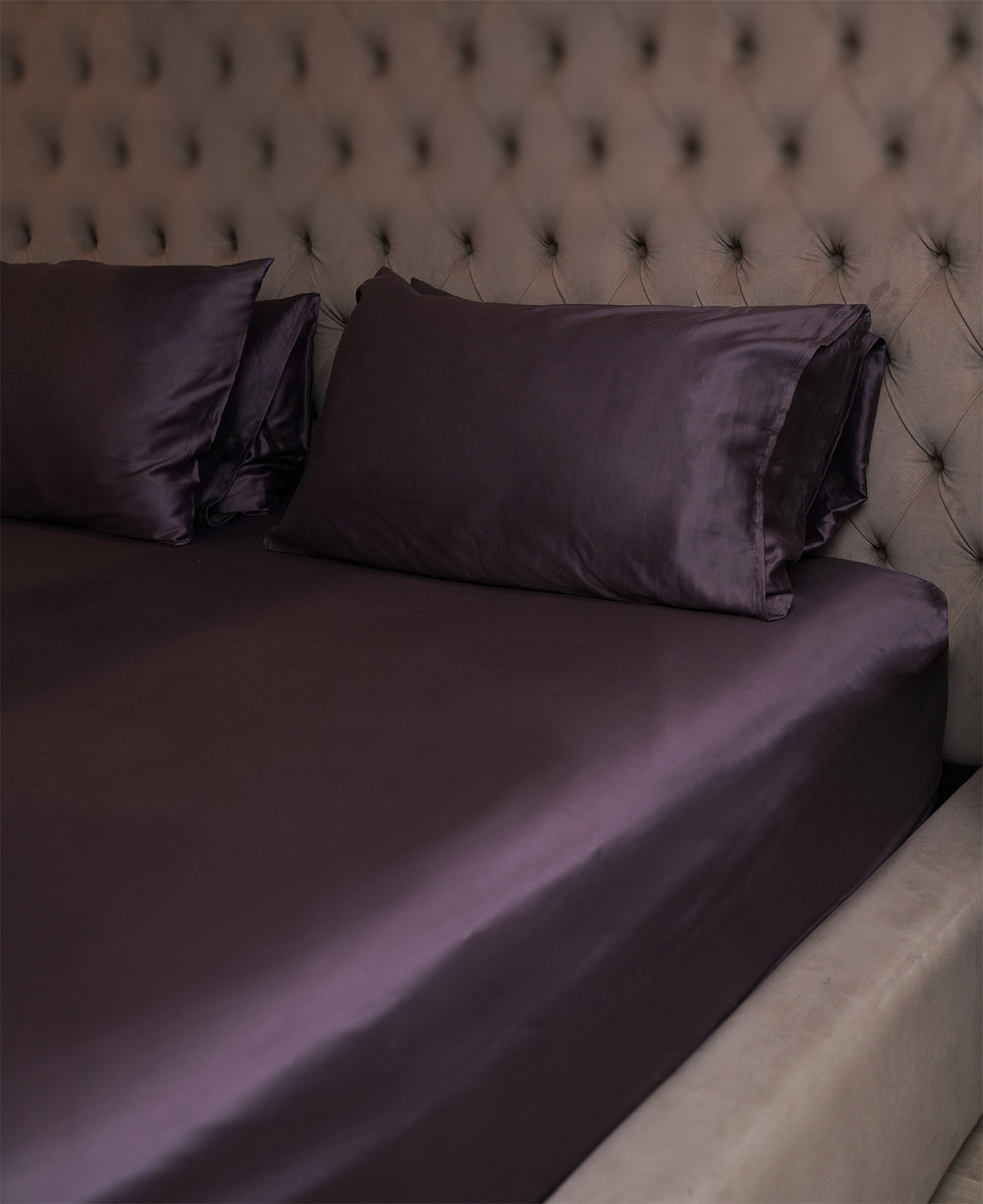 Purple satin sheets and pillows on a bed with a tufted headboard.